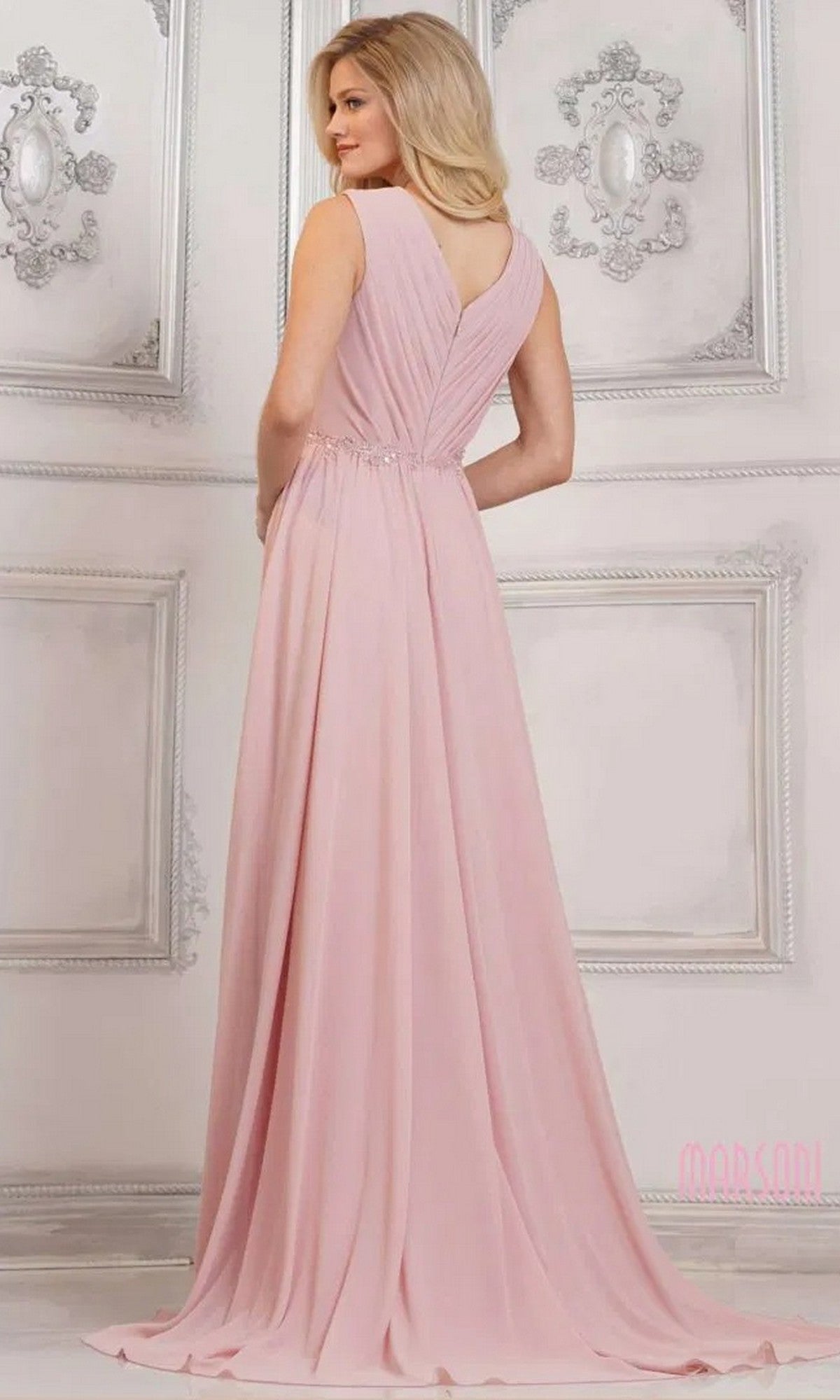 A woman stands in an elegant room wearing the Marsoni MV1292, a light pink, floor-length chiffon formal dress with a sleeveless design, V-shaped back, and beaded waist embellishments.